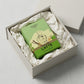 Angle : Vue de trois-quarts haut ($30^\circ$).Scène : Le carton de l'image image_888573.jpg est délicatement couché à l'intérieur d'un coffret de présentation e-commerce ouvert. Il est niché au milieu de papier de soie froissé d'un blanc crème impeccable. Deux petits flacons d'échantillons transparents sont discrètement posés dans un coin pour compléter la composition.Fond : Une surface de studio gris béton très clair et minimaliste. (gemini-3.1-flash-image-preview)