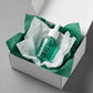 Angle : Vue oblique à mi-hauteur.  Scène : Le flacon d'Arencia est présenté à l'intérieur d'un coffret de livraison premium en carton blanc mat ouvert. Le produit est niché au milieu d'une superposition de papiers de soie froissés de deux couleurs : l'un blanc laiteux, l'autre vert émeraude pour rappeler le flacon.  Fond & Lumière : Surface de studio mate et contemporaine grise. Éclairage uniforme et très lumineux, mettant en valeur l'aspect e-commerce haut de gamme. (gemini-3.1-flash-image-preview)