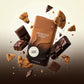 Une photo haute vitesse figée au format carré de 1200x1200px. Le flacon d'image_93c92b.jpg est le point focal absolu au centre de l'image. Autour de lui, des pépites de chocolat noir géométriques et des éclats de biscuits croquants flottent dynamiquement dans l'air, créant un effet de lévitation et de gourmandise brute. L'arrière-plan est un fondu doux allant du crème au brun foncé. (gemini-3-pro-image-preview)