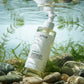 La caméra est positionnée bas, regardant à travers l'eau claire et ondulante. Le flacon d'Anua Cleansing Oil est à moitié submergé, reposant sur le lit de l'eau. Des plantes aquatiques et des algues délicates sont visibles sous la ligne de flottaison, soulignant la fraîcheur et la pureté. Le texte Anua est parfaitement clair. (gemini-3-pro-image-preview)