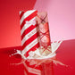 Angle : Vue oblique dynamique.  Scène : Le flacon et le carton de l'image image_7b5eae.jpg sont positionnés sur une surface laquée rouge ultra-brillante qui reflète leurs motifs à rayures. Une éclaboussure artistique de lait ou de crème blanche et liquide fige le mouvement autour de la base des produits, rappelant le côté crémeux du parfum.  Fond : Un dégradé de studio allant du rouge vif au rose pastel. (gemini-3-pro-image-preview)