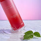 Angle : Face au flacon, au ras d'une surface liquide.  Scène : La base du parfum de l'image image_8c159e.png effleure à peine la surface d'une eau pure et transparente. Au moment de l'impact, de fines rides d'eau concentriques s'étendent. Une feuille de framboisier flotte à proximité.  Décor & Lumière : L'arrière-plan est un panneau de verre givré rétroéclairé par un dégradé rose et violet. Une lumière de studio ultra-rapide fige les éclaboussures microscopiques avec une netteté absolue. (gemini-3-pro-image