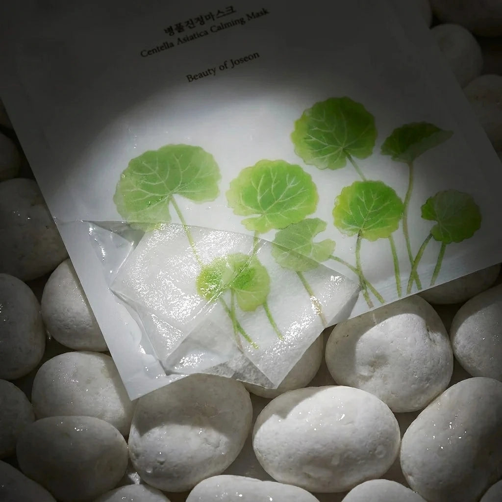 Angle : Gros plan serré sur la moitié inférieure du sachet.  Scène : Focus sur les illustrations de feuilles vertes et la fenêtre transparente. Le sachet est posé sur un lit de galets de rivière blancs et lisses, encore humides. La lumière révèle le contraste entre la rondeur minérale des galets et le pliage géométrique du masque en tissu gorgé de liquide à l'intérieur du sachet.  Décor & Lumière : Ambiance pénombre douce avec un spot ciblé sur le centre du produit pour faire briller le sérum en transparenc