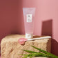 Angle : Face au produit (Eye-level shot), cadrage horizontal épuré, tube debout.  Scène : Le tube de l'image repose sur un bloc cubique de grès ou de travertin beige très clair, poreux et texturé. Une fine spatule en céramique blanche est posée sur la pierre rugueuse juste devant le packaging, une noisette généreuse de gel rose/translucide brillante s'étirant depuis son extrémité. Quelques feuilles de riz vert éclatant sont disposées à la base pour un contraste de couleur complémentaire.  Éclairage : Fond m